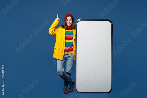 Full body young woman in sweater red hat yellow raincoat big huge blank screen area mobile cell phone point finger up pisolated on plain dark blue background Outdoors wet fall weather season concept photo