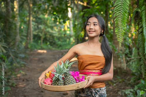 Young girl in traditional dress with a bowl full of tropical fruits walking through the tropical forest