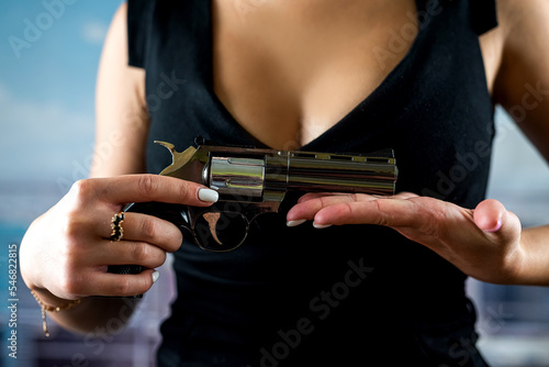 faceless woman in stylish dress holds revolver for shot against background of office windows.