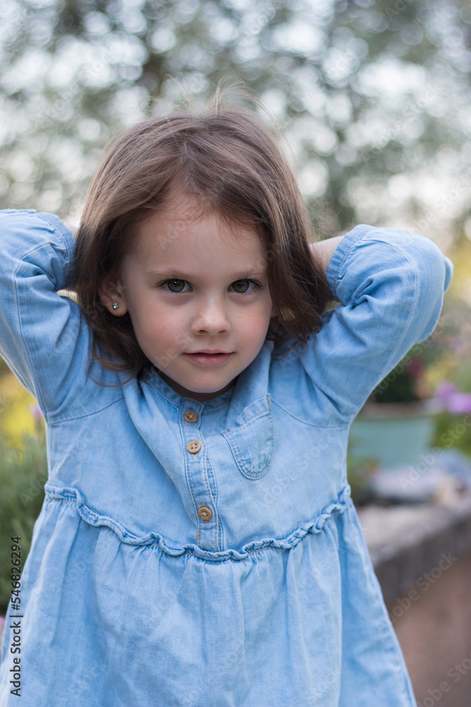 A little beautiful preschooler girl stands in a denim blue dress against the background of flowers A little beautiful preschooler girl stands in a denim blue dress against the background of flowers