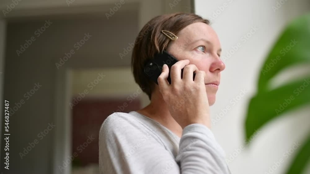Sad alone woman talking on mobile phone while looking out the window of living room. Casual mid-adult brunette with serious facial expression Sad alone woman talking on mobile phone while looking out the window of living room. Casual mid-adult brunette with serious facial expression