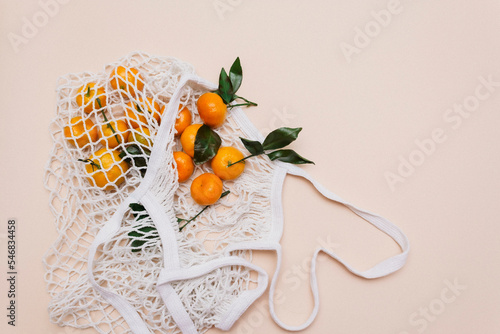 Top view of a white grocery eco-mesh cotton bag with fresh orange tangerines on a beige pastel background. Healthy food, zero waste, healthy lifestyle