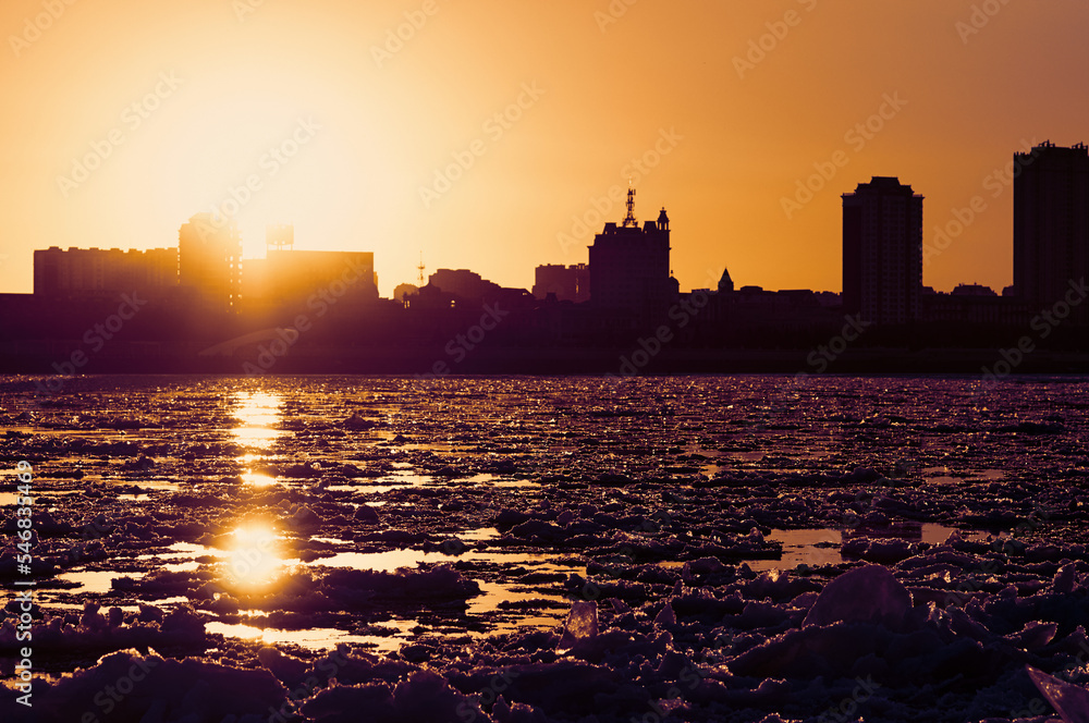 Sunset on the river bank covered with ice floes during ice drift. Silhouettes of city buildings in the sun. Glare on water and ice floes. Embossed surface texture. Photography in Low key.
