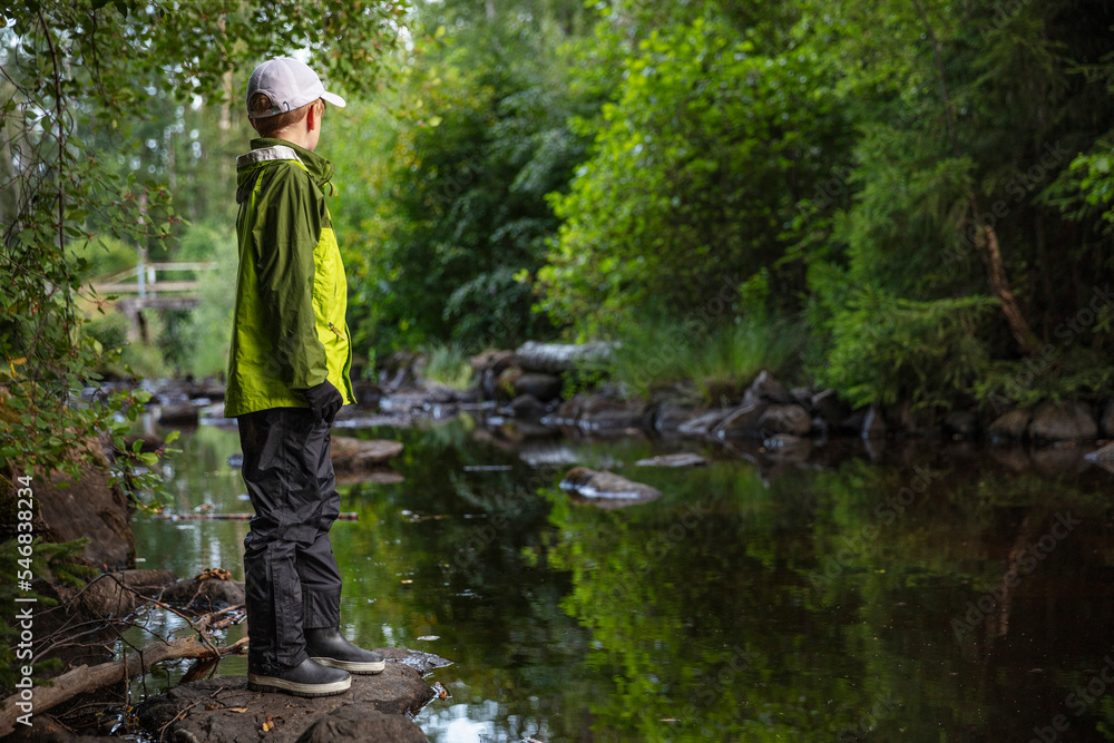 Boy in cap and jacket standing on rock by stream Boy in cap and jacket standing on rock by stream