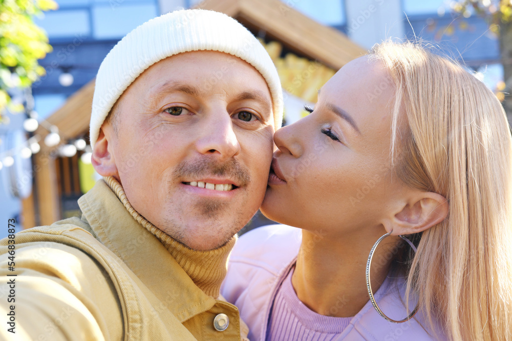 A beautiful young couple is looking at the phone camera, smiling and taking a selfie. Blonde girl kissing a guy on the cheek