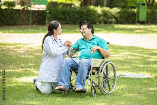 Indian caregiver nurse taking care of senior male patient in a wheelchair outdoor at park, Asian doctor help and support elderly mature older people. rehabilitation and health care.