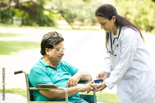 Indian doctor check with pulse oximeter device on finger of senior female diabetes patient in a wheelchair outdoor at park.