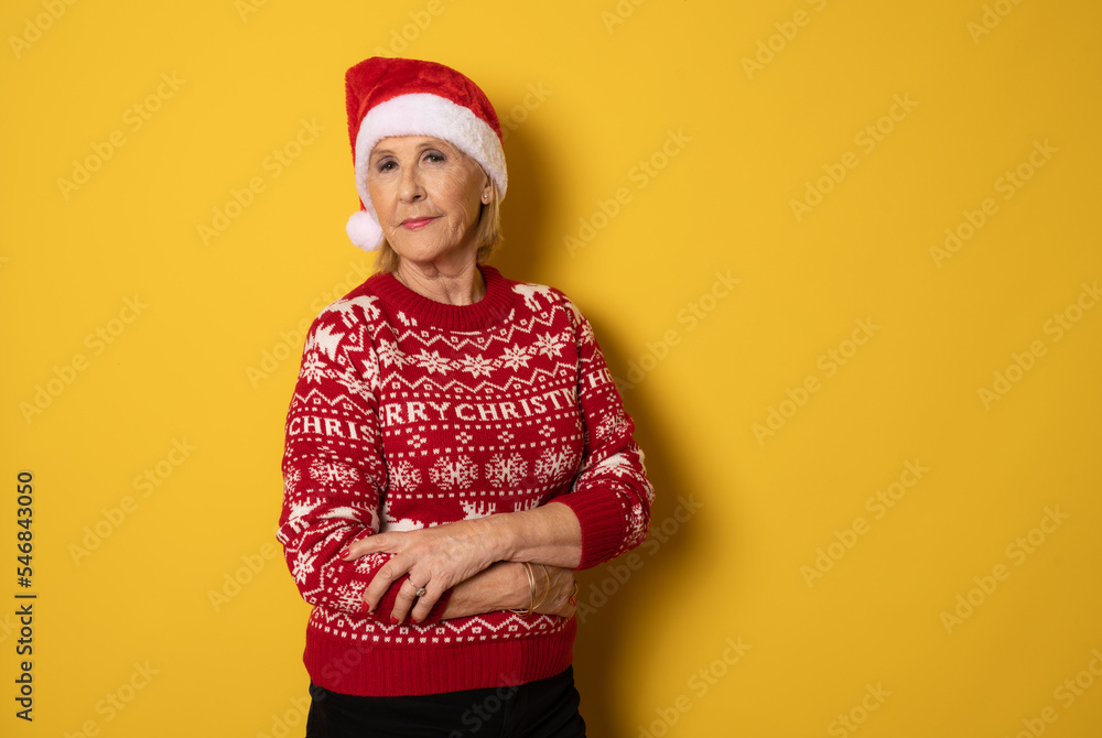 Happy confident senior woman wearing red santa hat stands with arms crossed and looking at the camera isolated on yellow. Mature lady promoting new year deal, holiday sale Happy confident senior woman wearing red santa hat stands with arms crossed and looking at the camera isolated on yellow. Mature lady promoting new year deal, holiday sale