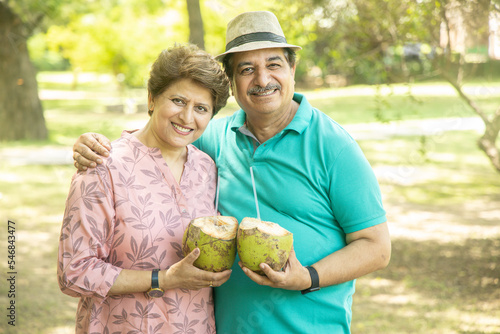 Happy indian senior couple having coconut water at summer park. Old people enjoying retirement life.