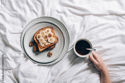 Open sandwich from slice of wholegrain bread with peanut nut butter, bananas and crushed walnuts. Breakfast in bed with plate and coffee on bedding