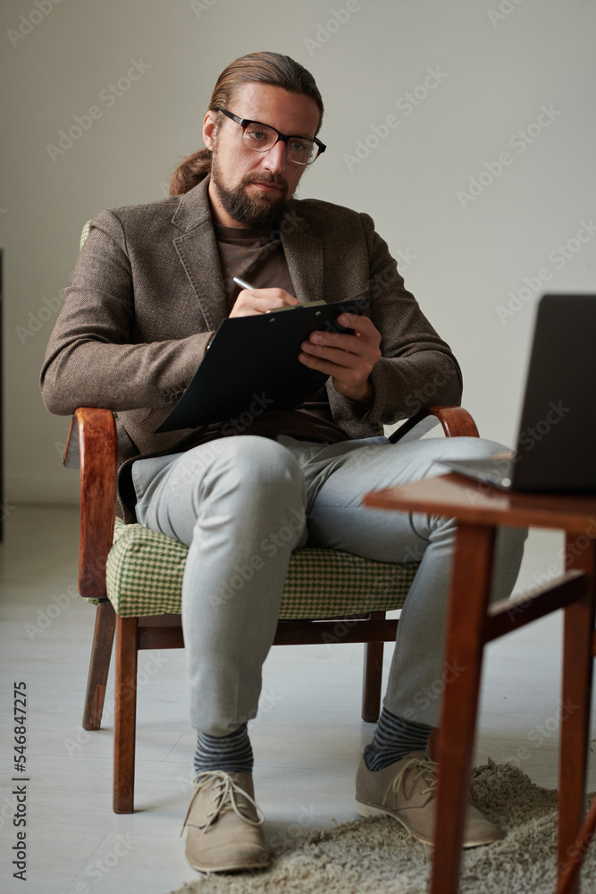 Serious psychologist having online meeting on laptop at office, he sitting on armchair and making notes in document