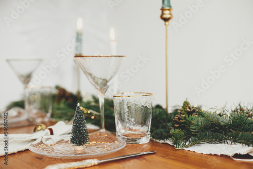 Stylish Christmas table setting. Christmas little tree on plate, linen napkin with bell, vintage cutlery, wineglass, fir branches with golden lights, candles on table. Atmospheric Holiday brunch