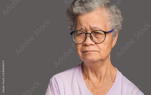 An elderly Asian woman is looking at the camera with a serious while standing on a gray background