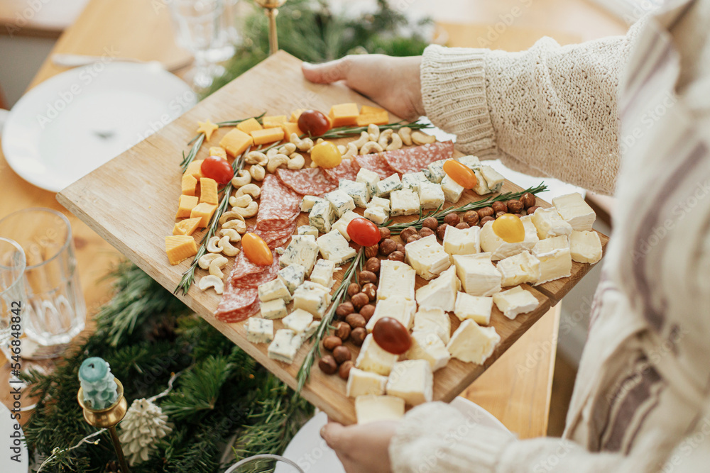 Woman holding cheese board on background of stylish christmas table with fir branches and candles. Cheese appetizers and salami in shape of christmas tree, creative food arrangement for holidays