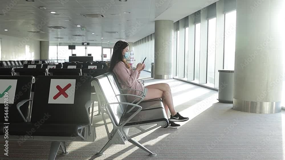 A young woman wearing a mask at the airport during the Covid-19 pandemic sits in a chair in an empty departure lounge. social distance A young woman wearing a mask at the airport during the Covid-19 pandemic sits in a chair in an empty departure lounge. social distance