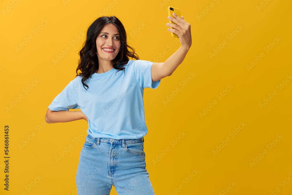 Woman blogger with a phone in her hands in a blue t-shirt and jeans on a yellow background smile signs gestures symbols, online communication and video call, copy space, free background Woman blogger with a phone in her hands in a blue t-shirt and jeans on a yellow background smile signs gestures symbols, online communication and video call, copy space, free background