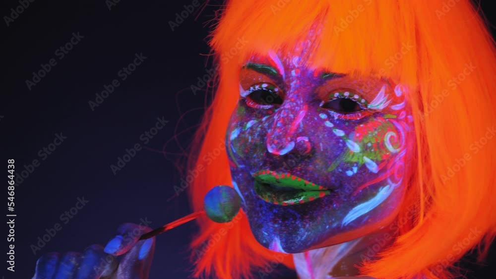 Close-up of a girl with an ultraviolet body art and an orange wig eating a Lollipop in the dark under fluorescent lights. Body art glowing in UV light. Close-up of a girl with an ultraviolet body art and an orange wig eating a Lollipop in the dark under fluorescent lights. Body art glowing in UV light.