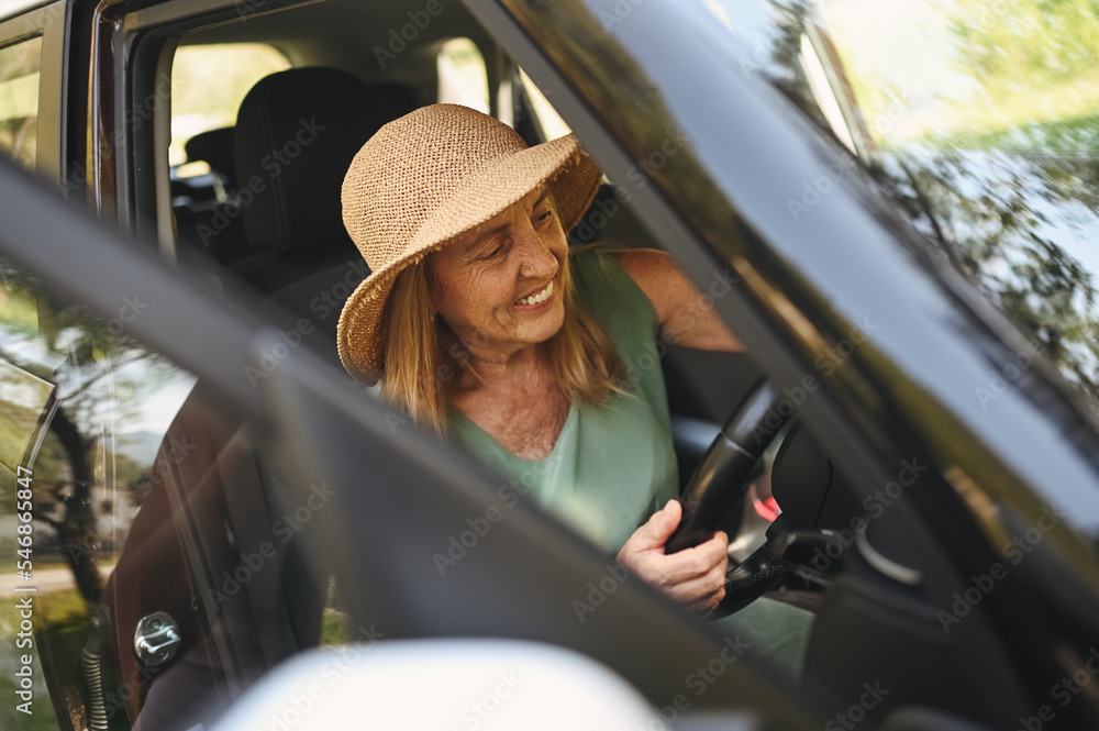 Naklejka premium Happy senior woman driver in straw hat driving sitting in new car, smiling looking at camera enjoying journey. Driving courses and life insurance. Retired people activity and road trip concept 