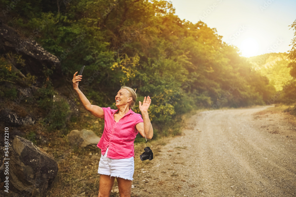 Happy senior woman tourist walking in summer forest road outdoors at sunset time. Old slim lady traveling with photo camera makes selfie on smartphone. Active retirement vacation concept. Warm filter