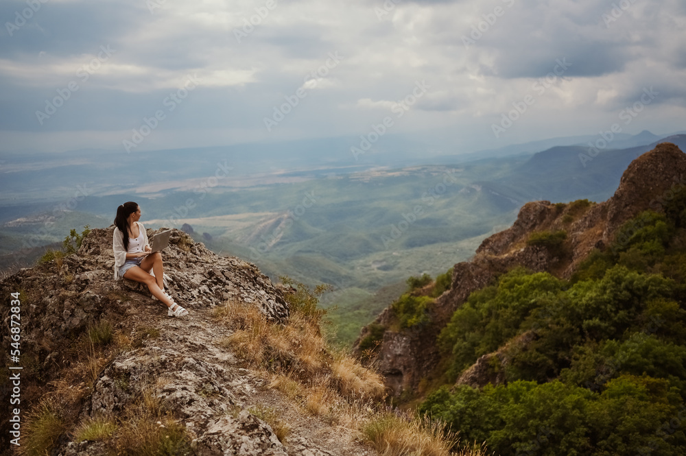 Young woman freelancer traveler working online anywhere outdoors using laptop enjoying mountain peak view. Happy female downshifter holding computer in unusual amazing view workplace at summer