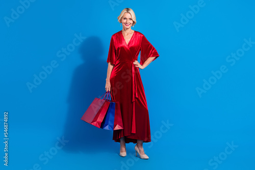 Full length photo of charming cute senior lady wear red shiny dress holding bargains isolated blue color background
