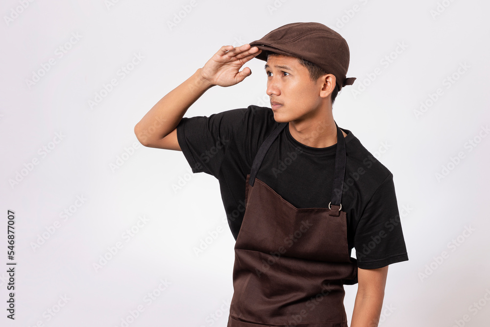 Handsome barista asian man wearing brown apron and black t-shirt isolated over white background looking far away at distance with hand over head. Vision concept. Handsome barista asian man wearing brown apron and black t-shirt isolated over white background looking far away at distance with hand over head. Vision concept.