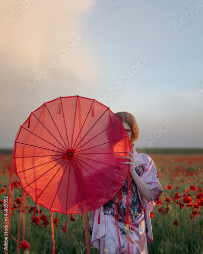 A girl in a kimono hides behind an umbrella with ribbons in a field of poppies