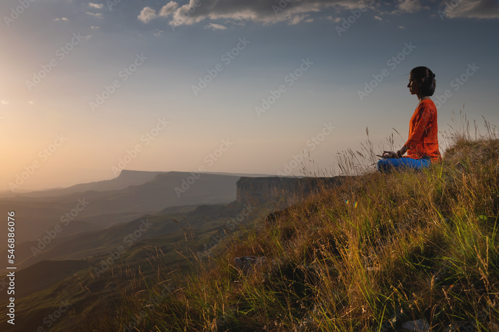 meditating woman sitting in lotus position near a cliff at sunset overlooking the mountains, practice in yoga, peace of mind
