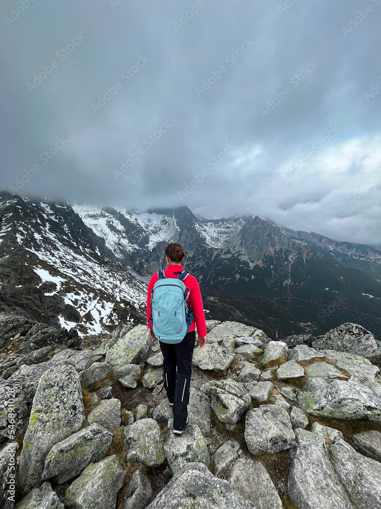 girl in red hoody and with blue backpack is hiking in snowy mountains at the end of autumn, beginning of winter. view from back, active life concept. Vertical, grey sky