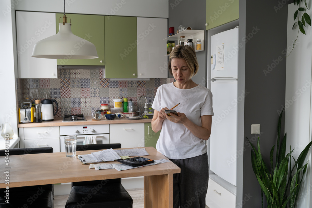 Busy woman with bunch of unpaid bills on table tries to solve problems with obligations standing in kitchen. Middle aged female wants to pay bills searching contacts of hotline on Internet on phone