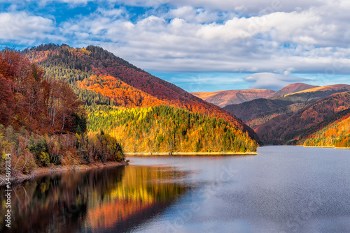 Scenic view of autumn colored forest reflecting to alpine lake in Transylvanian Alps during warm October light against dramatic sky