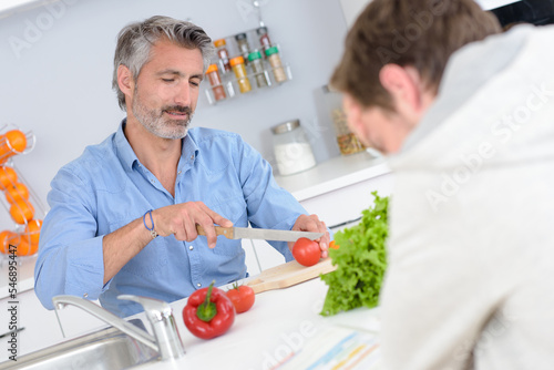 Man chopping vegetables while another man reads