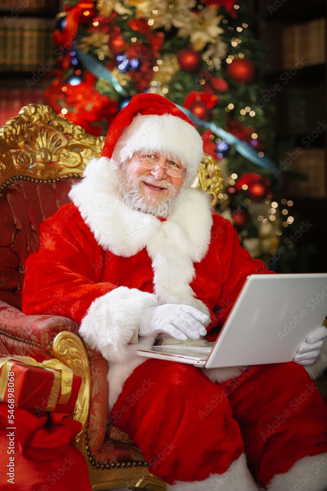 Santa Claus using a laptop computer. Sitting in a chair next to the decorated Christmas tree, chatting with friends. Santa Claus using a laptop computer. Sitting in a chair next to the decorated Christmas tree, chatting with friends.