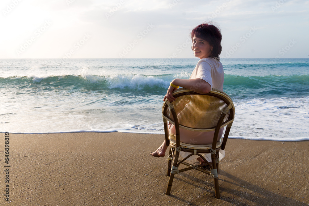 An adult woman on the beach is sitting on a chair looking at the sea waves.