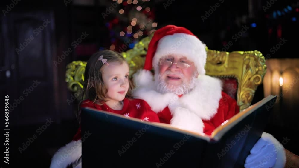 Portrait of a happy Santa Claus sitting at home with a little assistant reading a Christmas wish book Portrait of a happy Santa Claus sitting at home with a little assistant reading a Christmas wish book