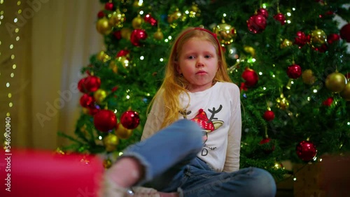 A sad girl is sitting next to a Christmas tree who received an unwanted gift on Christmas Day