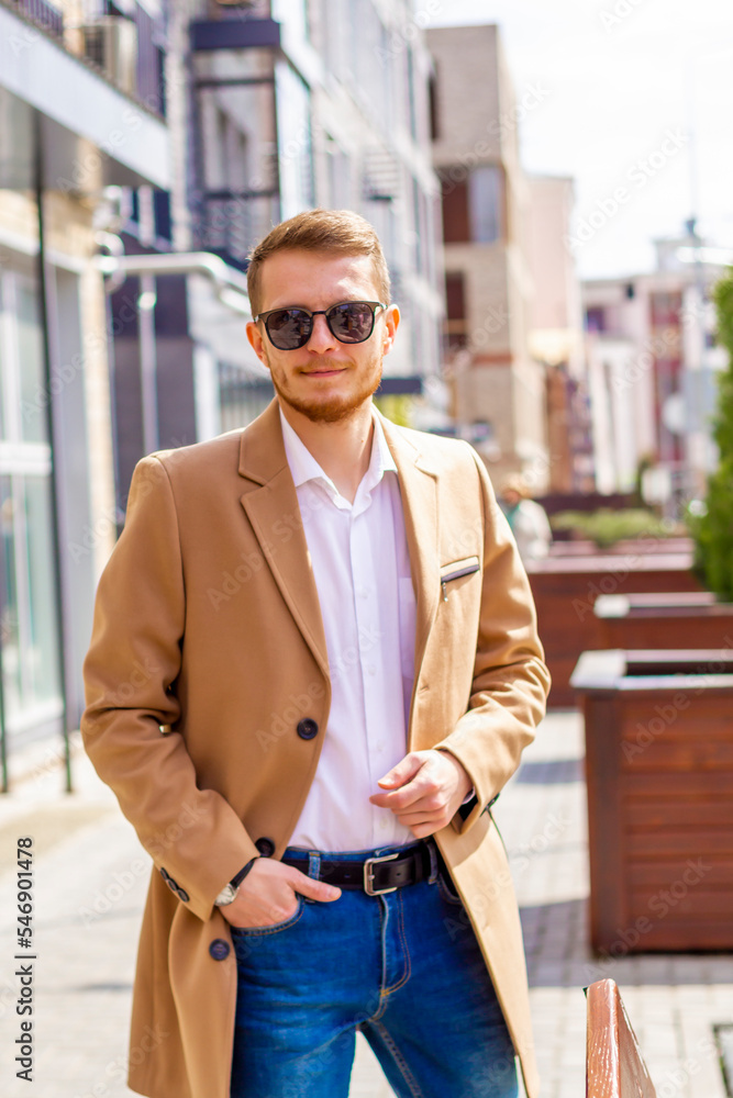 A young man dressed in a beige coat is walking down the street. A young bearded guy with a modern hairstyle and sunglasses on an urban background