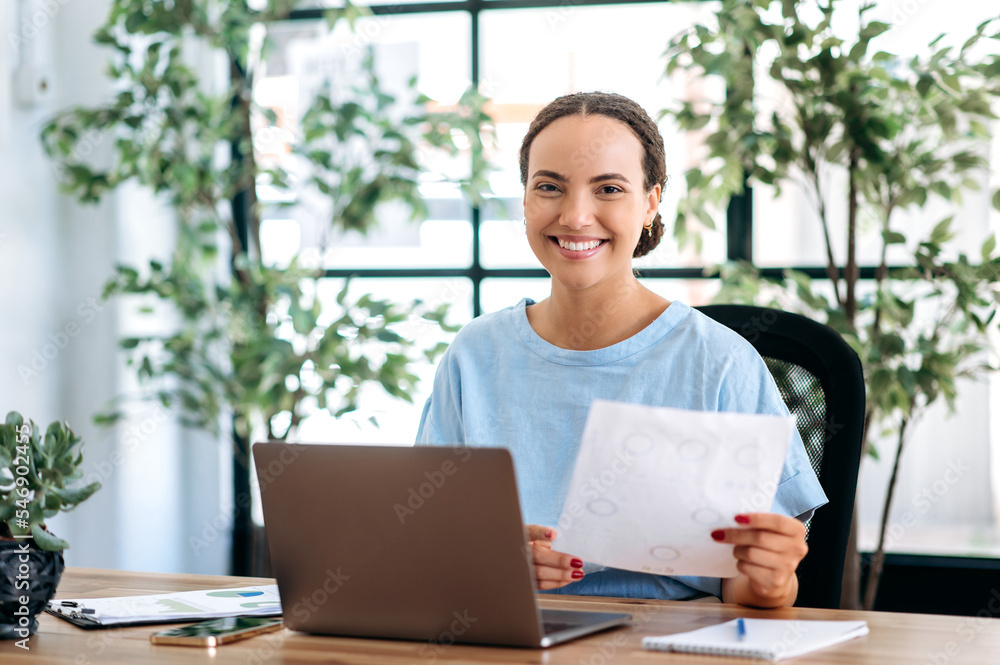 Successful intelligent mixed race business woman, financial ceo, analyst, sits at a work desk in a modern office, holds financial documents in her hands, looks at the camera, smiles happily Successful intelligent mixed race business woman, financial ceo, analyst, sits at a work desk in a modern office, holds financial documents in her hands, looks at the camera, smiles happily