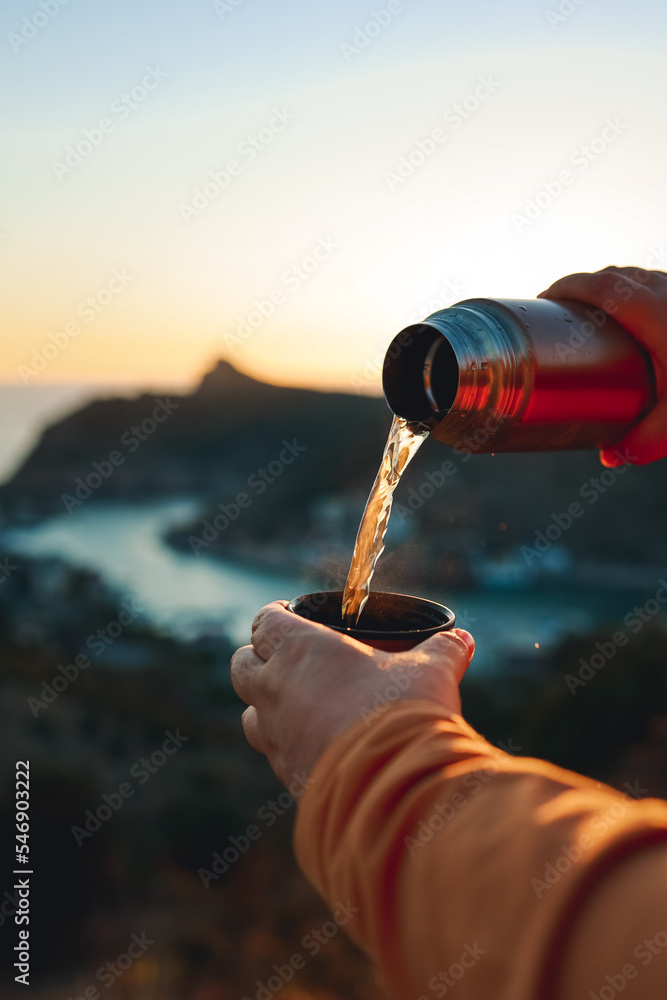 Naklejka premium A human pours hot tea from a thermos into a mug on the mountain. Concept of autumn outdoor recreation. 
