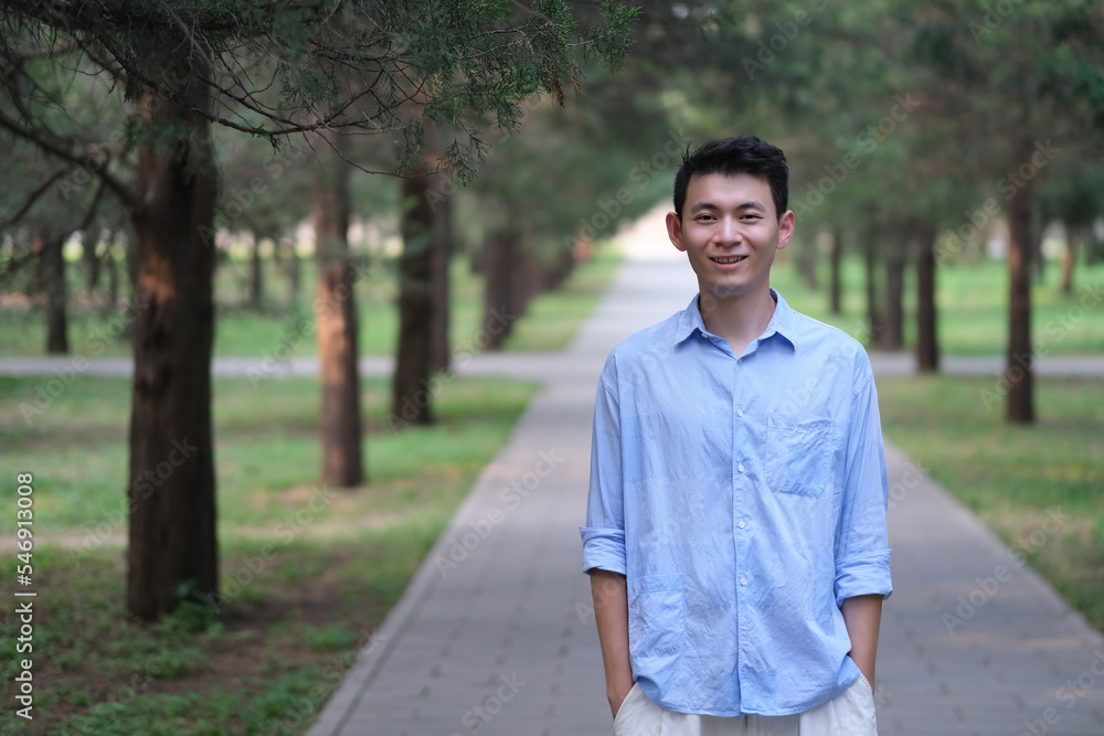 smiling handsome Asian young man, hands in pockets, at green park path, looking at camera smiling handsome Asian young man, hands in pockets, at green park path, looking at camera