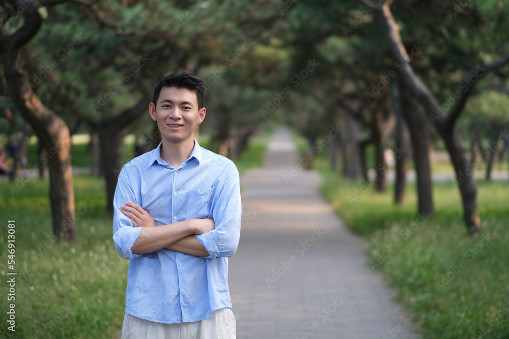 smiling handsome Asian young man, cross arms, at green park path, looking at camera smiling handsome Asian young man, cross arms, at green park path, looking at camera