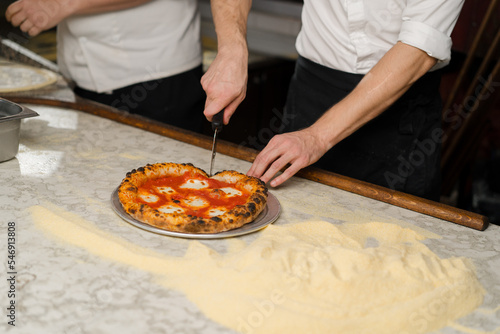 A professional chef cuts Margherita pizza with a knife in the restaurant kitchen. Traditional Italian cuisine