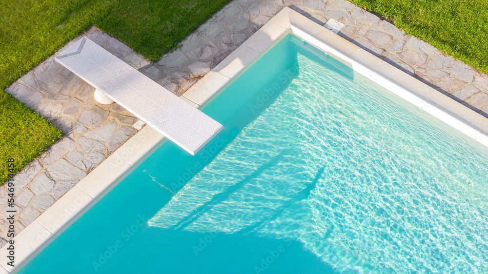 Aerial view of a rectangular swimming pool with diving board, belonging to a large villa. The water is transparent and through the blue you can see the steps. Around the water there is a stone floor.