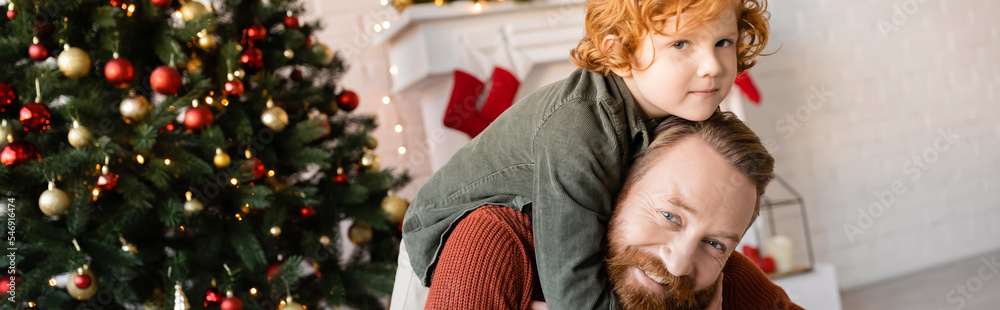happy bearded man piggybacking redhead son near blurred Christmas tree, banner happy bearded man piggybacking redhead son near blurred Christmas tree, banner
