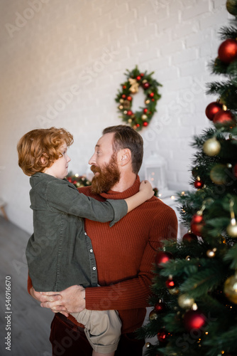 cheerful bearded man holding redhead son in hands near blurred pine tree decorated with Christmas baubles