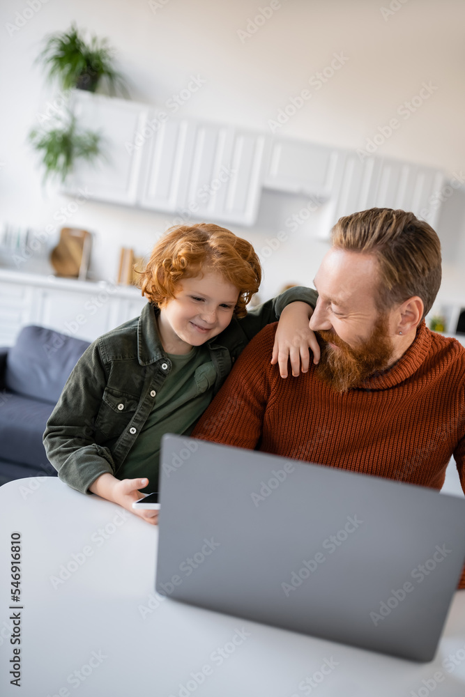 happy boy standing near smiling father working on blurred laptop at home happy boy standing near smiling father working on blurred laptop at home