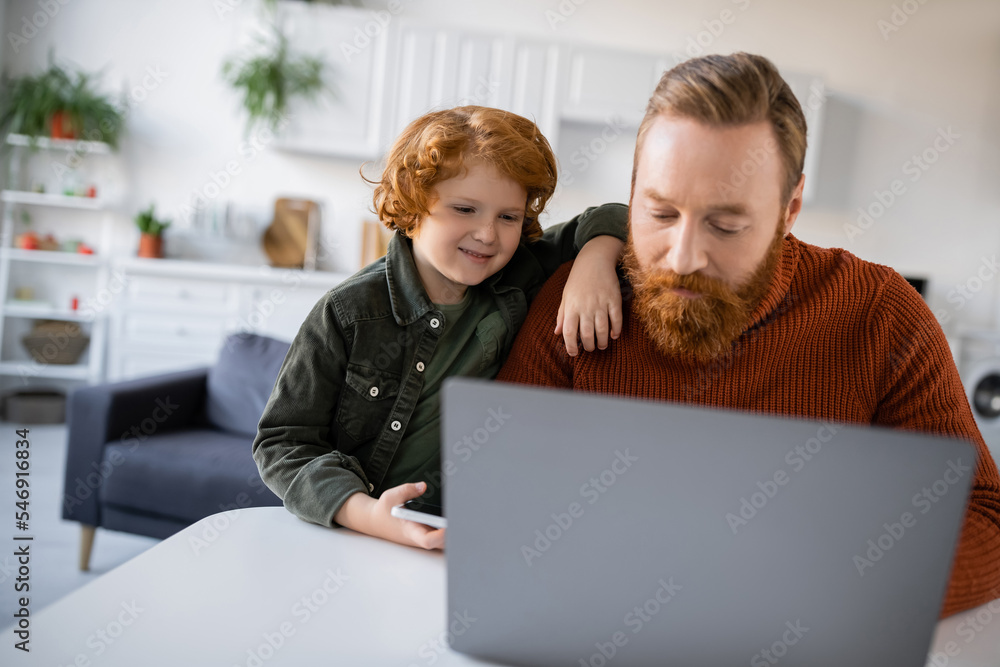 redhead kid smiling near bearded dad working on blurred laptop at home redhead kid smiling near bearded dad working on blurred laptop at home