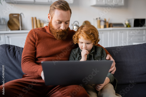 thoughtful redhead child looking at laptop while sitting on couch near bearded dad
