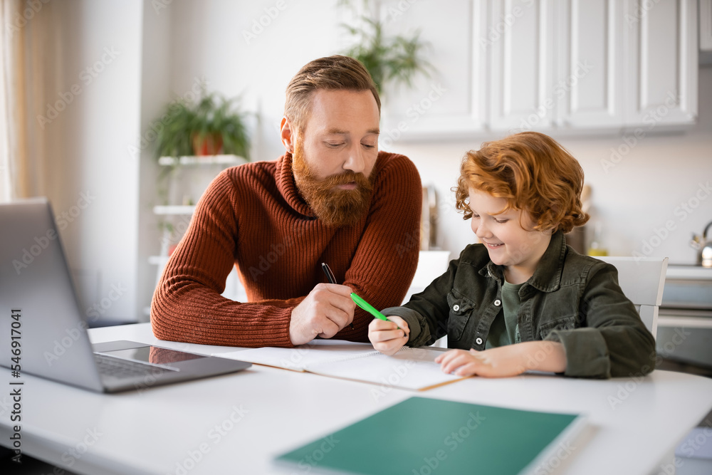 Fototapeta premium bearded man assisting smiling redhead son writing in notebook near blurred laptop