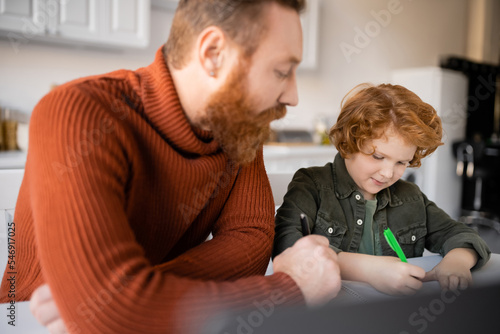 bearded man looking at redhead son writing in notebook while doing homework on blurred foreground